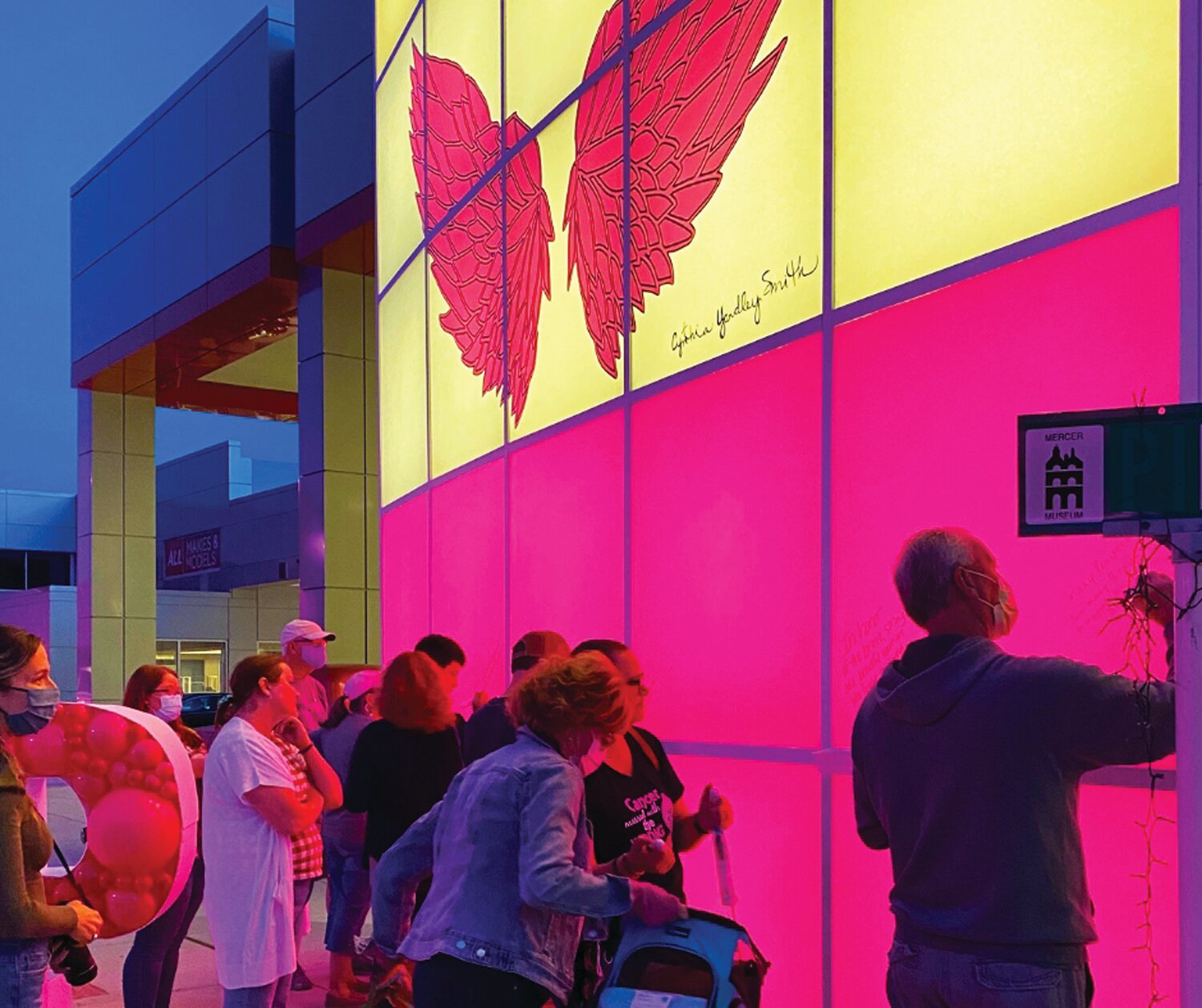 Community members signing the luminous pink panels on the front of the Thompson Toyota dealership, symbolizing unity and support for breast cancer survivors and those in recovery.