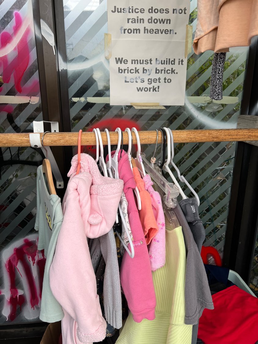 Full text on the flyer taped above this rack of children’s clothes reads, “Justice does not rain down from heaven. We must build it brick by brick. Let’s get to work!” (Photo by Jill Boogren)