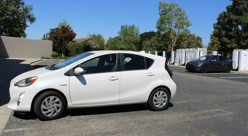 A white Toyota Prius C, a hybrid, is parked near an electric vehicle charging at a Tesla station.