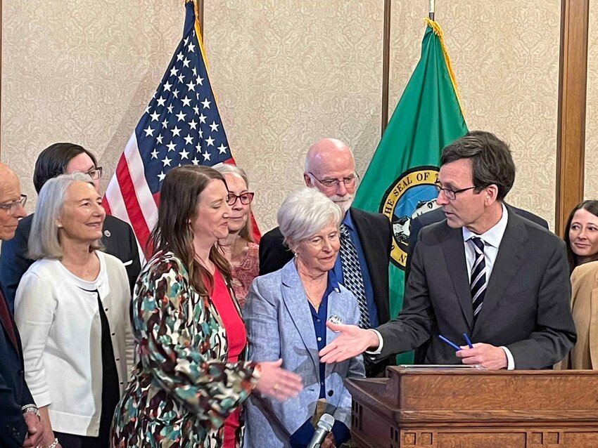 Gov. Bob Ferguson, at podium, goes to shake hands with state Sen. Noel Frame, D-Seattle, at the signing of a bill to make clergy mandatory reporters of child abuse and neglect, on May 2, 2025 in Olympia. Standing between them is Mary Dispenza, a founding member of the Catholic Accountability Project. (Photo by Jerry Cornfield/Washington State Standard)