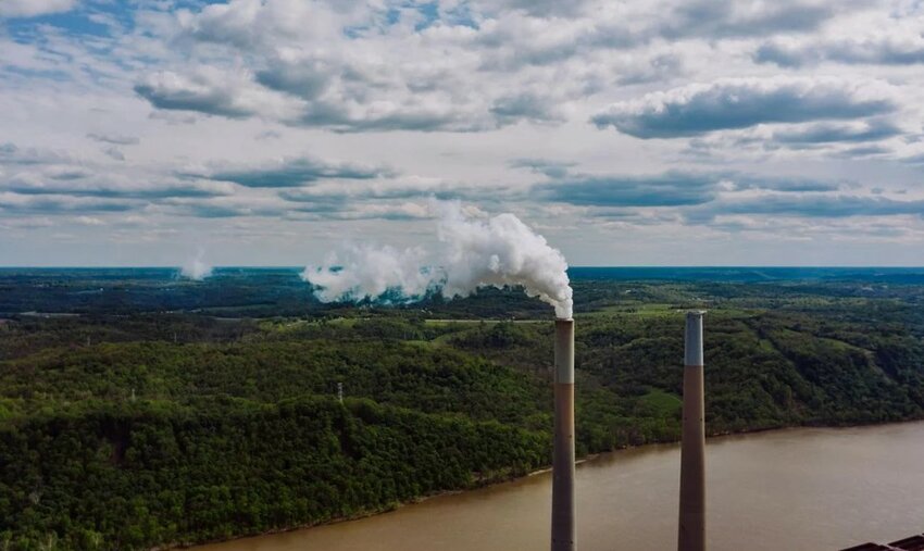 Two smokestacks emit plumes over a river and forested valley.