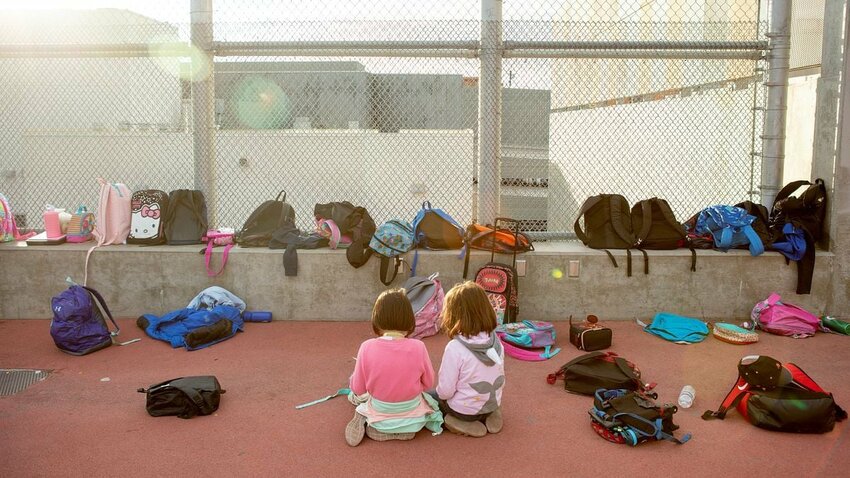 Two young children sit together on a schoolyard surface surrounded by colorful backpacks.